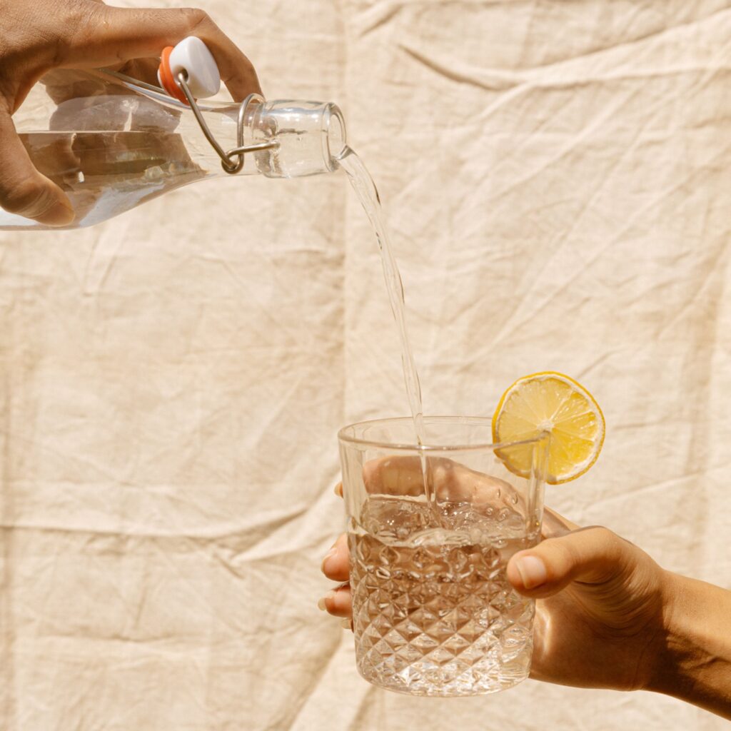One hand holds a clear glass filled with water and a side of lemon while a second hand pours from a jug of water