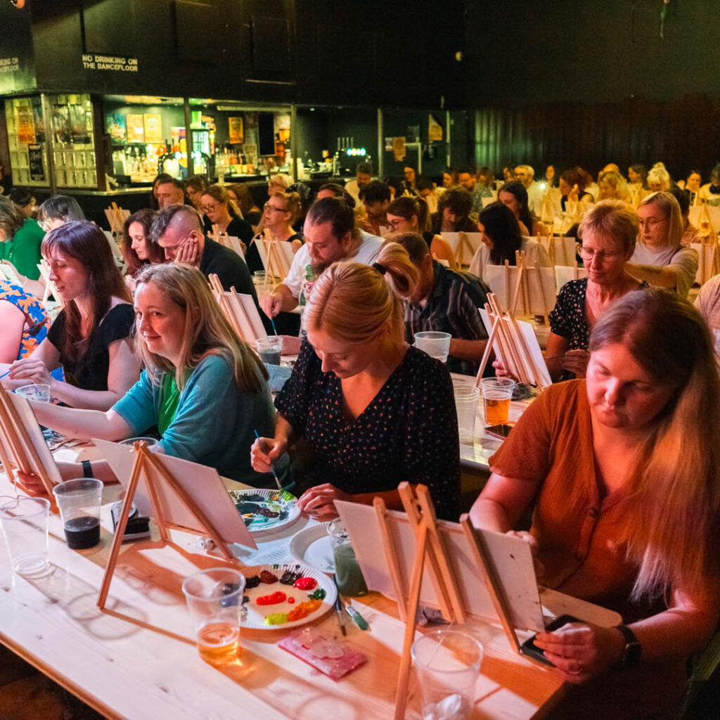 Rows of people paint on mini canvases during a Bob Ross Paint Along event at Digital Newcastle.