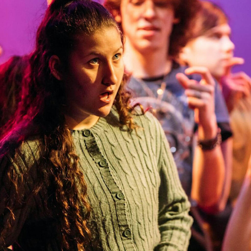 A young woman in a green jumper opens her mouth and looks at the ceiling as she performs in the show Brainstorm. Other young people can be seen making gestures in the background.