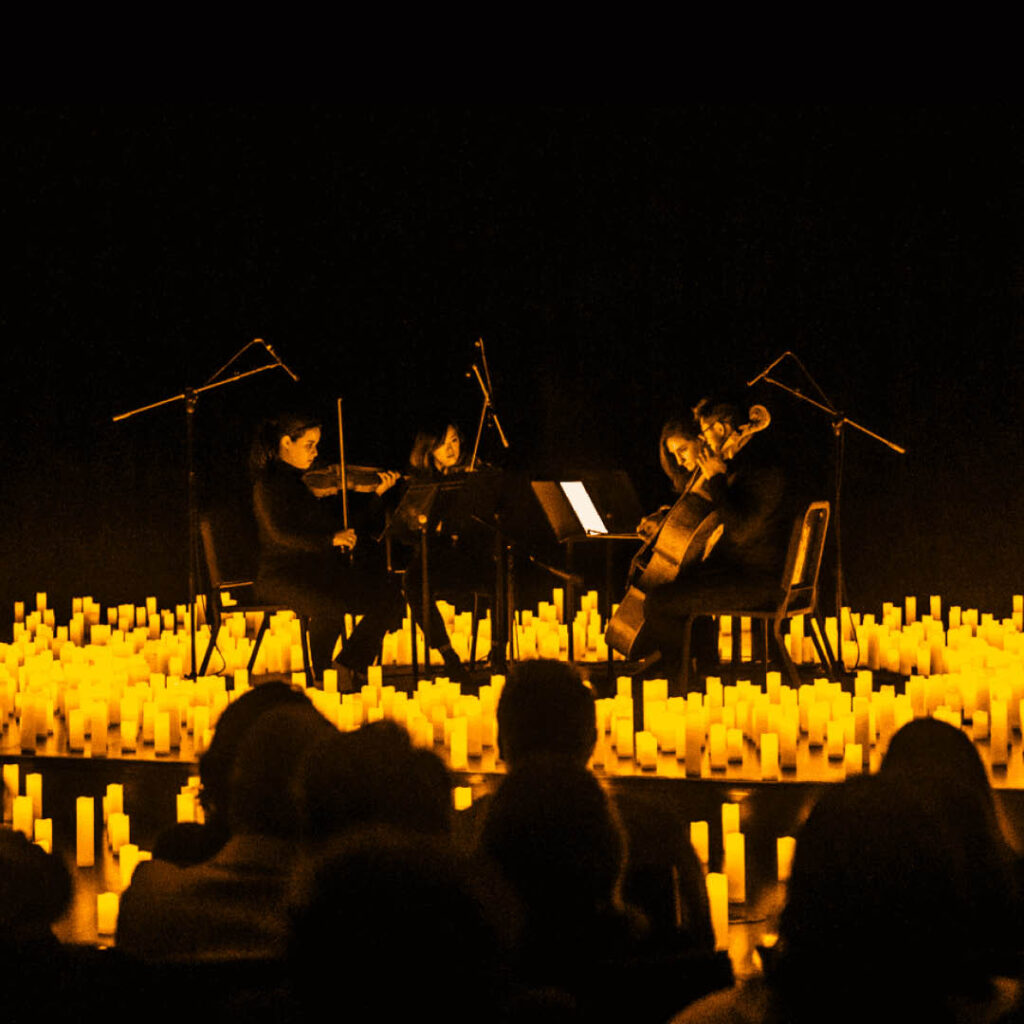 Four string players sit in a circle on a stage covered with lit candles. They concentrate on playing their music. In the foreground, the silhouettes of the audience can be seen.