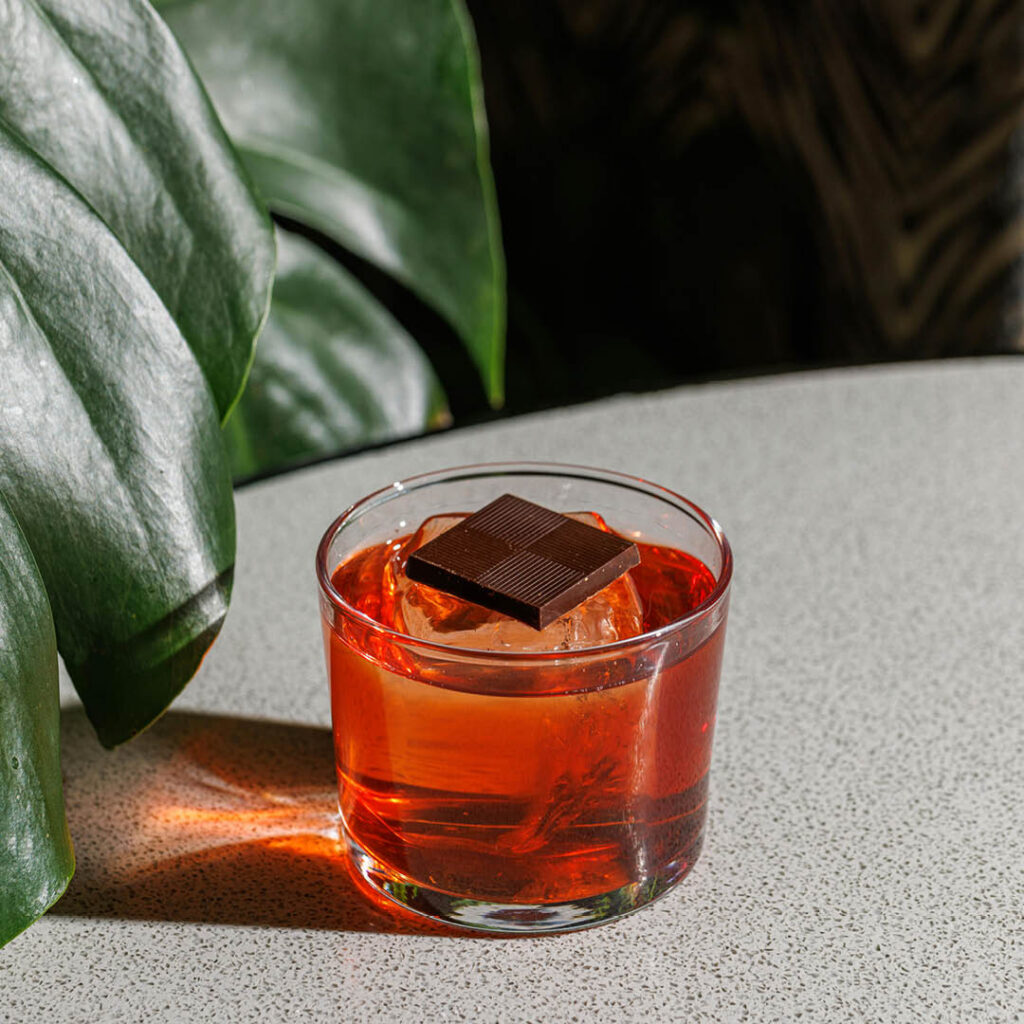 A red cocktail in a tumbler glass sits on a grey marbled surface next to a large plant frond. It is topped with a square of dark chocolate.