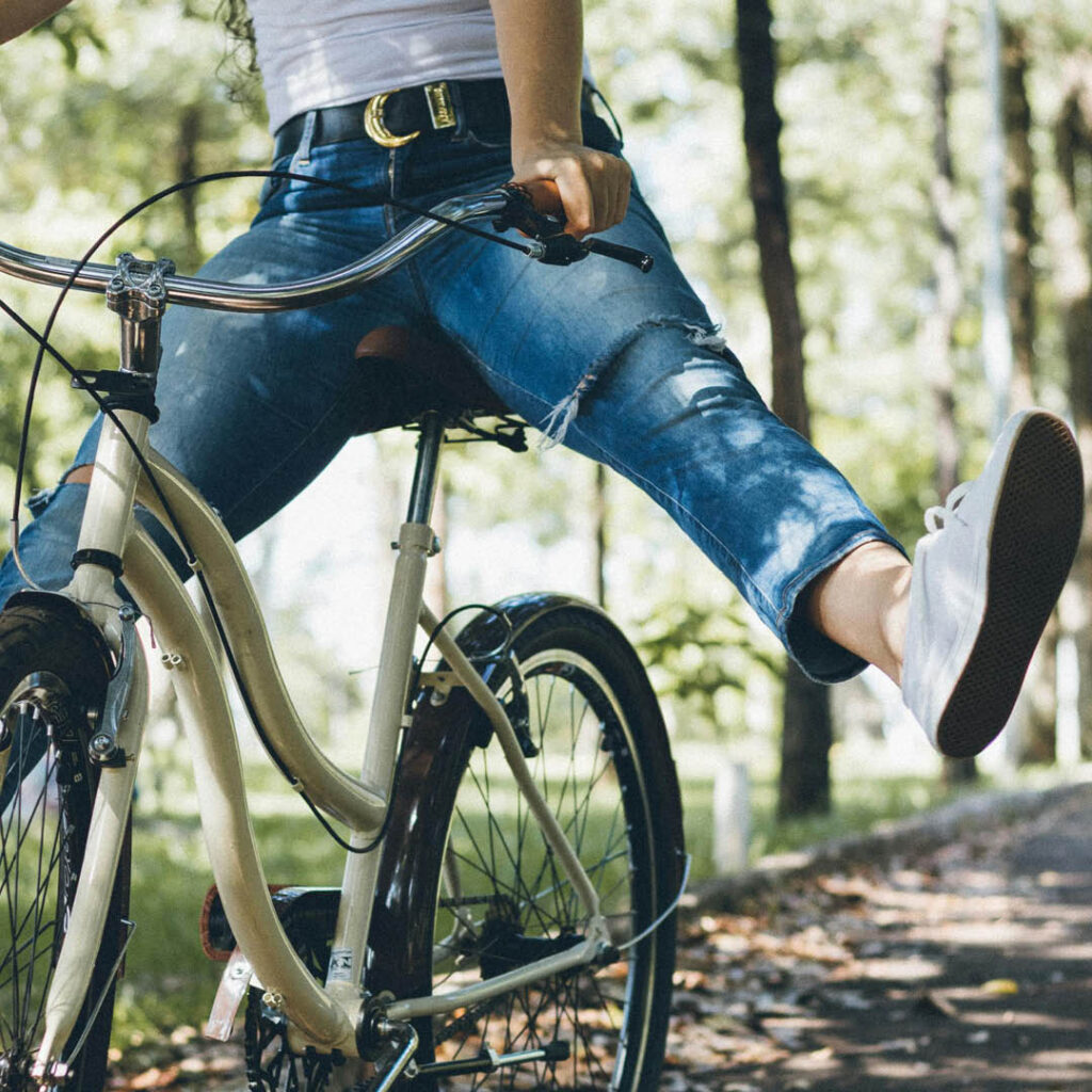 A person rides a bike along a park path. They raise both of their legs off the pedals as they glide along in the sun.