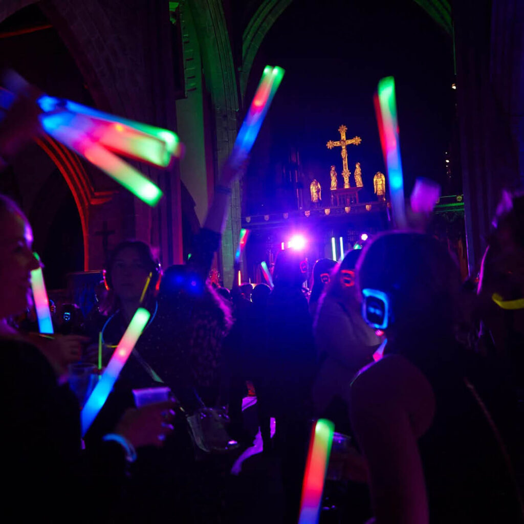Crowds of people hold up multi coloured glow sticks in the dark abbey. A golden cross glows at the front of the room above the dancing silhouettes.