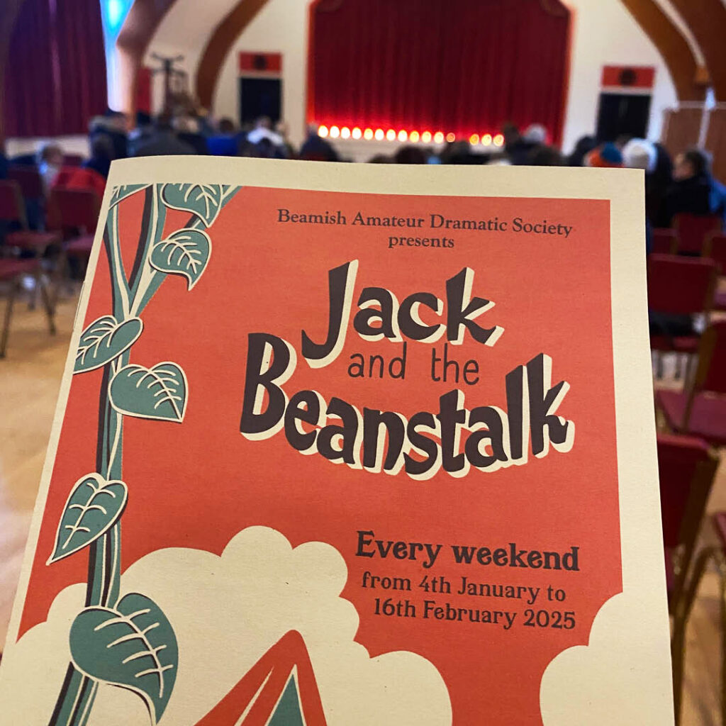 An unseen person holds up a red paper programme for the Beamish Jack and the Beanstalk pantomime. In the background, there is a red curtained stage and a people sitting on rows of chairs in front of it.