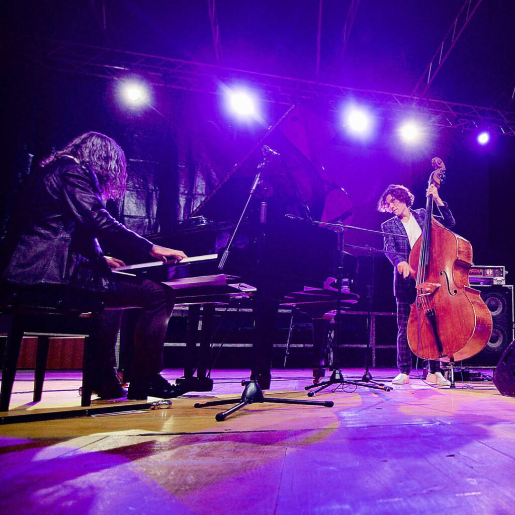 A man stands and plays the piano on a purple lit stage. He looks at another man sitting at a piano, who has long black hair which hangs forward as he plays.