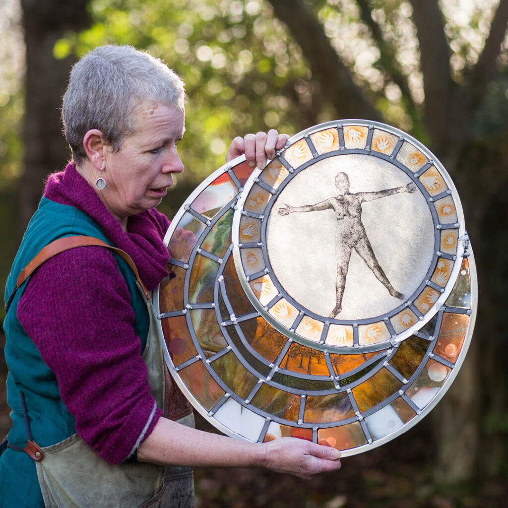 Glass artist Helen Grierson looks at her artwork as she holds it up outside. Circular plates of glass feature different coloured pieces. The top plate features the shape of a man.