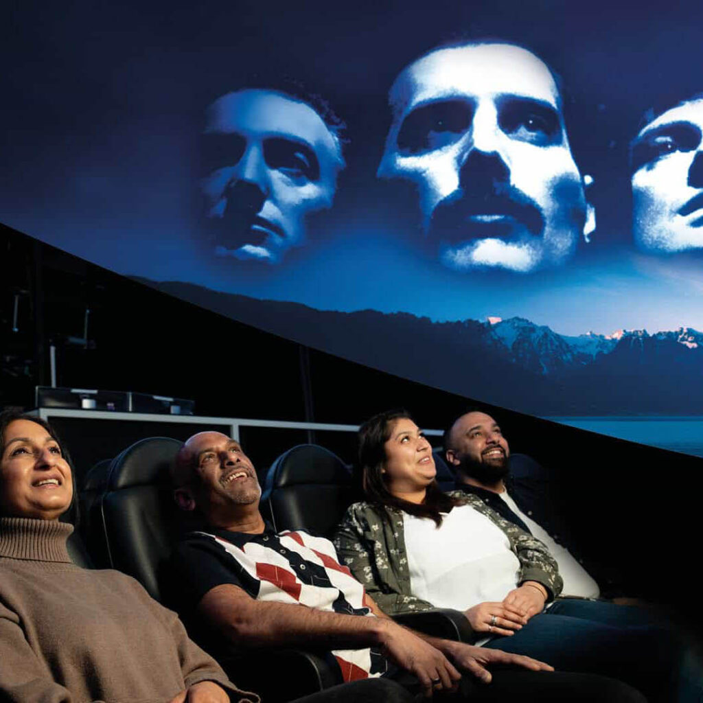 Four adults smiles as they lie under a planetarium and gaze up at the projected faces of Queen on the domed ceiling above them. They are at Life Science Centre in Newcastle.