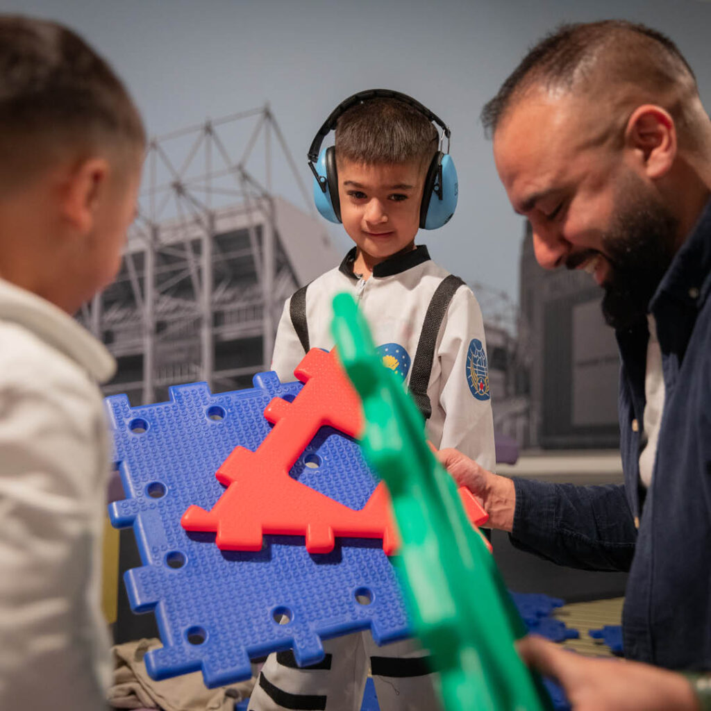 A little boy wearing noise cancelling headphones looks in interest as his dad holds up some colourful building pieces. He is enjoying a relaxed Sunday session at Life Science Centre.