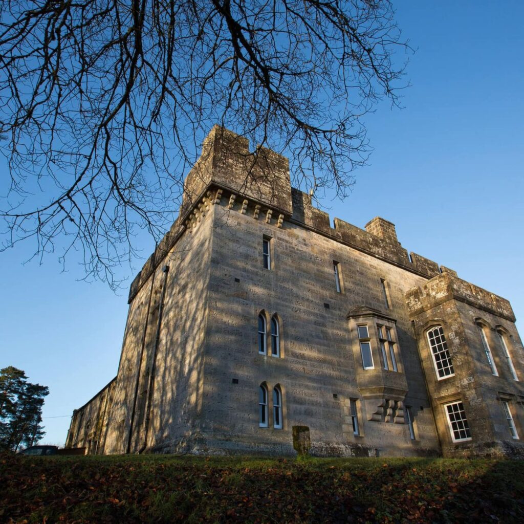 A big historic Kielder castle is seen through some tree branches against a blue sky