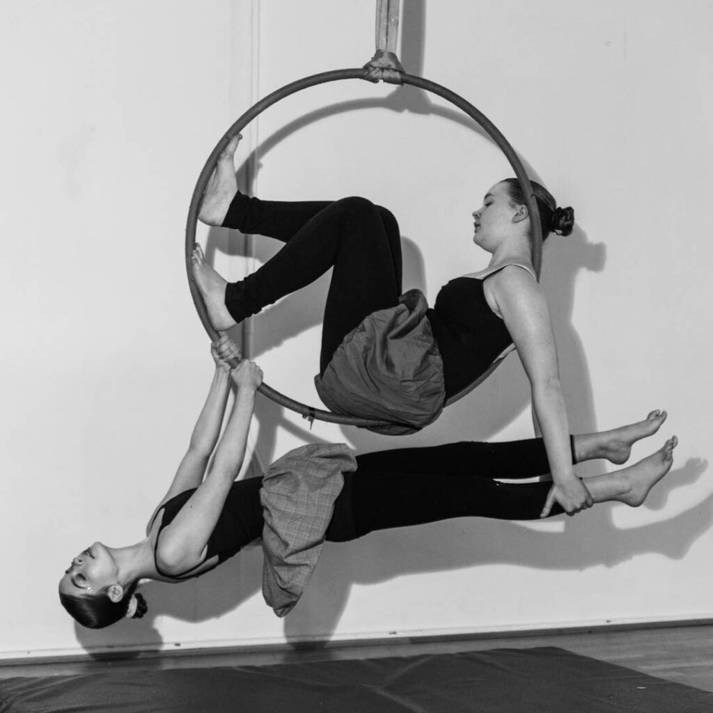 Two girls pose during an aerial hoop workshop. One of them sits in a suspended hoop, holding the legs of the girl hanging below her.