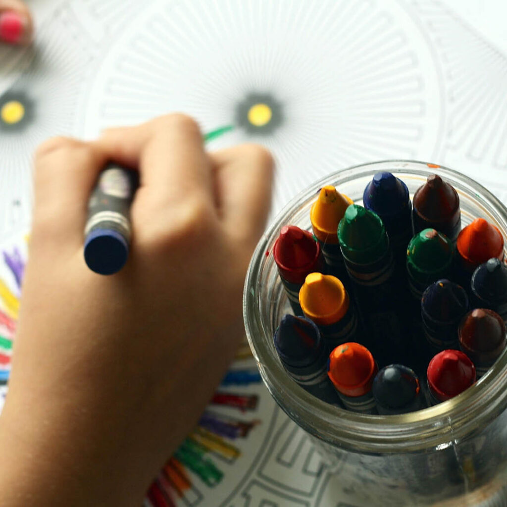 An unseen child uses a crayon to colour in a mandala colouring sheet. A pot of various coloured crayons stand on top of the sheet of paper.