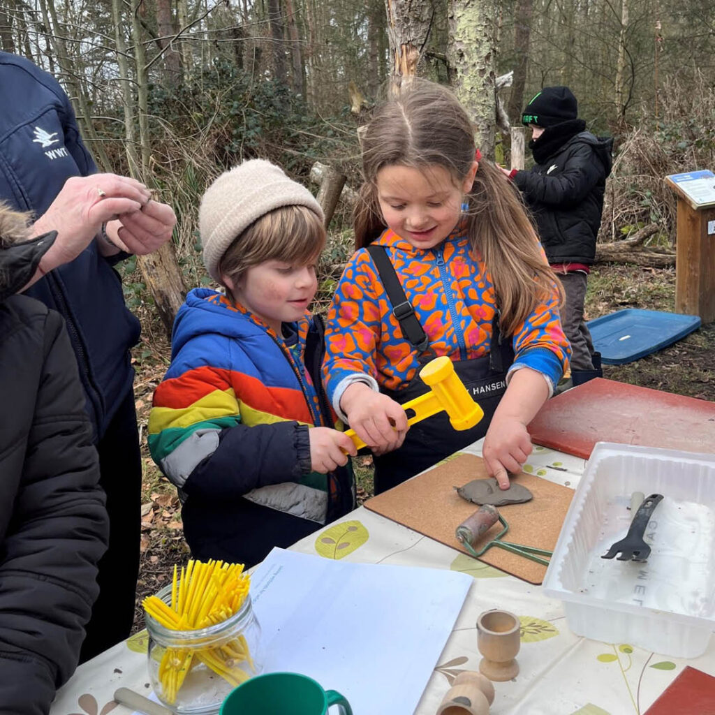 A little boy and girl stand in the woods in front of a craft table. Their faces are muddy and they smile as they hammer a piece of clay into shape.