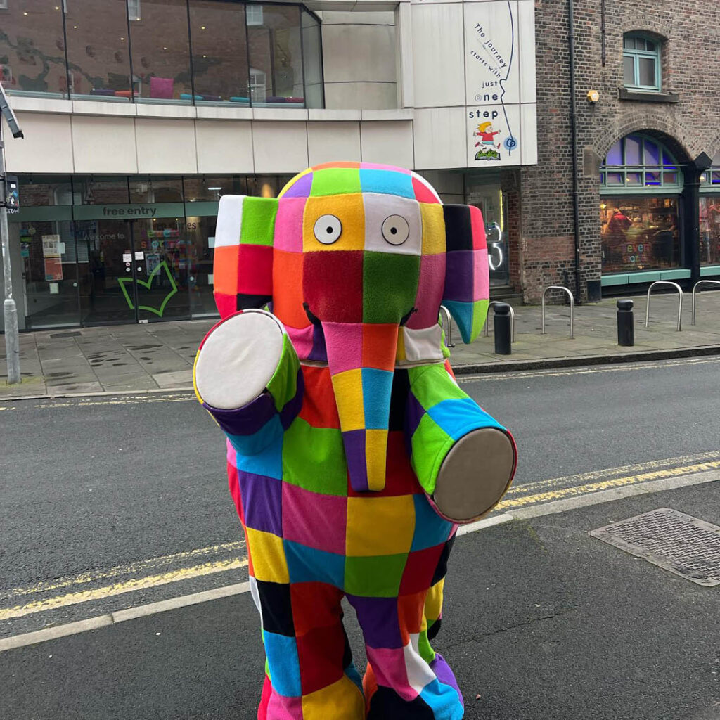 A person dressed in a colourful Elmer the Elephant costume stands outside of Seven Stories. They hold their arms up in welcome.
