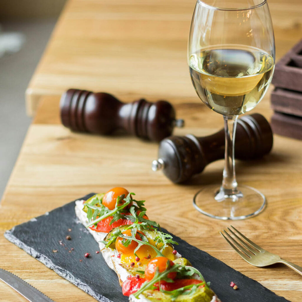 A glass of white wine stands next to a long piece of bruschetta.