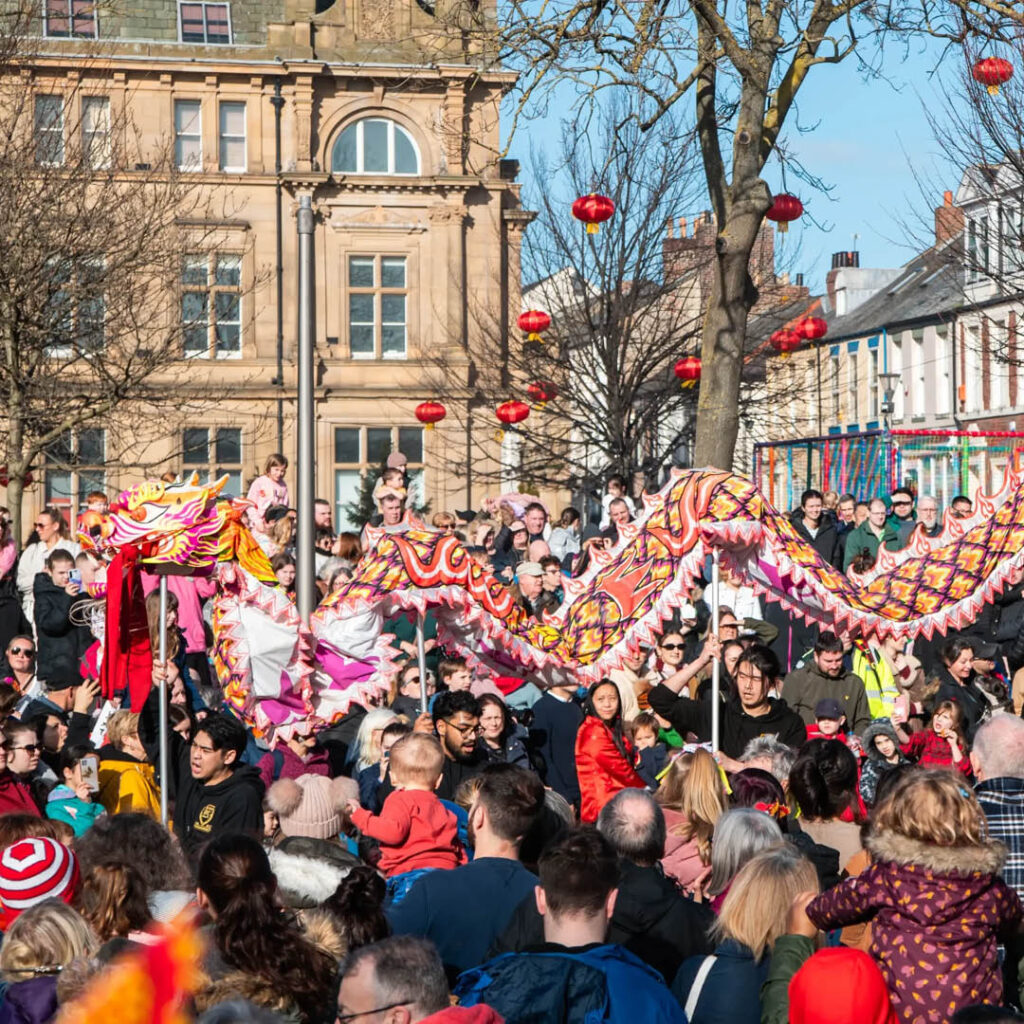 A crowd of people gather outside on a sunny day. A large Chinese dragon winds its way through the crowd. Red Chinese lanterns hang in the nearby trees. This is part of the Lunar New Year celebrations at Sunniside Gardens in Sunderland.