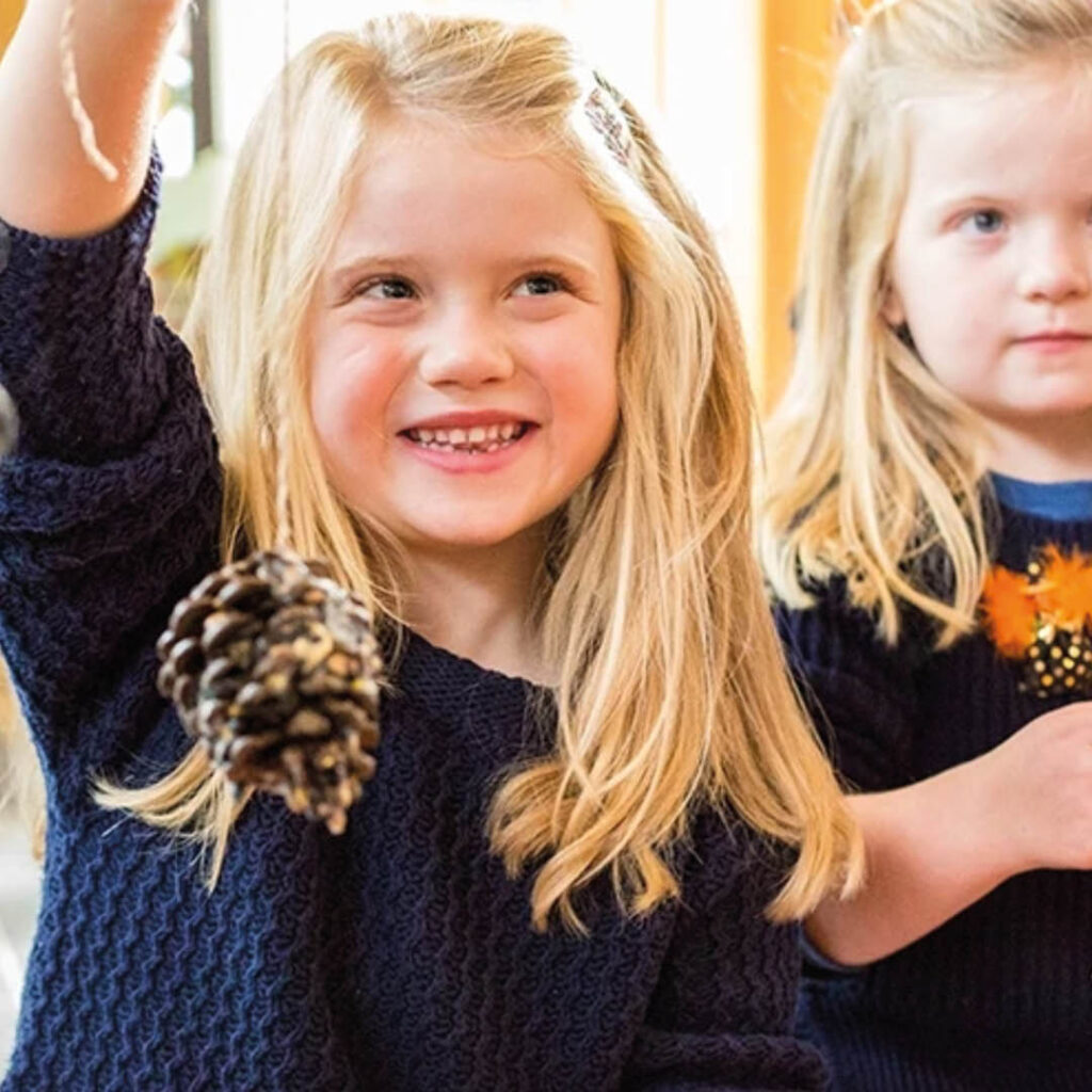 A little girl with long blonde hair grins as she holds up a bird feeder made from a pine cone.