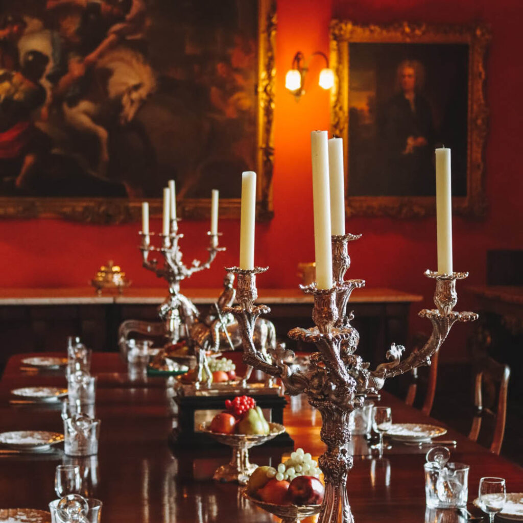 Red walled dining room. The long wooden dining table is laid with bowls of fruit and silver candelabras holding white candlesticks. It is the dining room at Raby Castle.