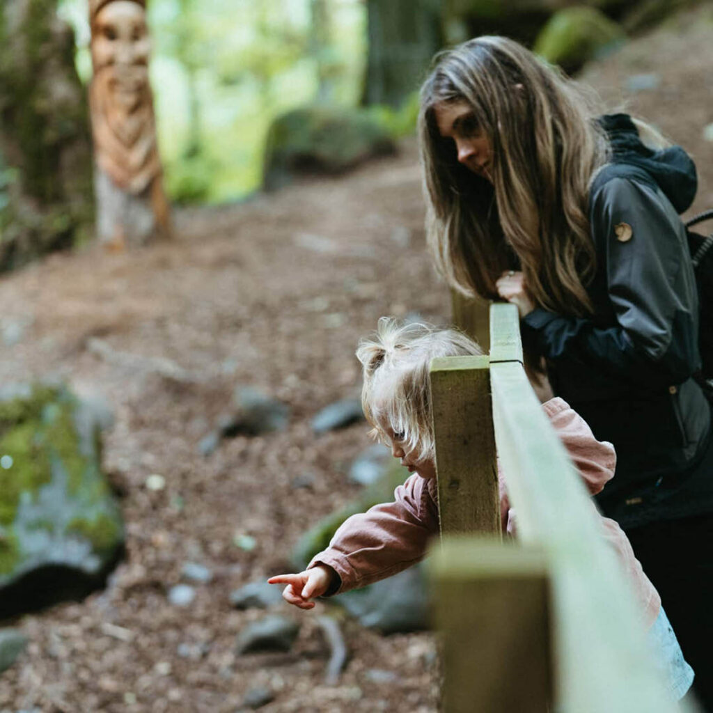 A mother and her young daughter stand in the woodland at High Force Waterfall. The little girl leans through a wooden fence and points at something in the distance while the mother looks on.