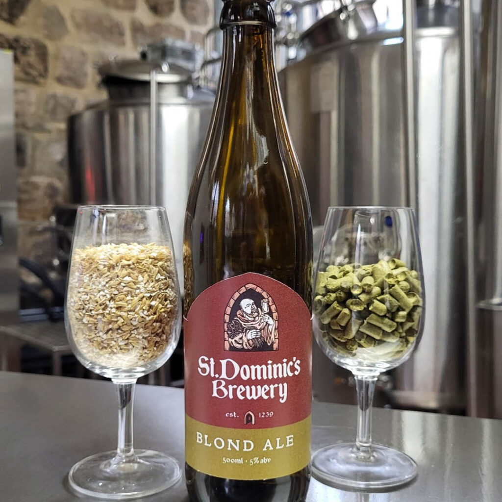 A bottle of St Dominics Brewery beer sits on a stainless steel surface in front of brewery apparatus. There is a pint glass on either side, each filled with hops or brewing pellets.