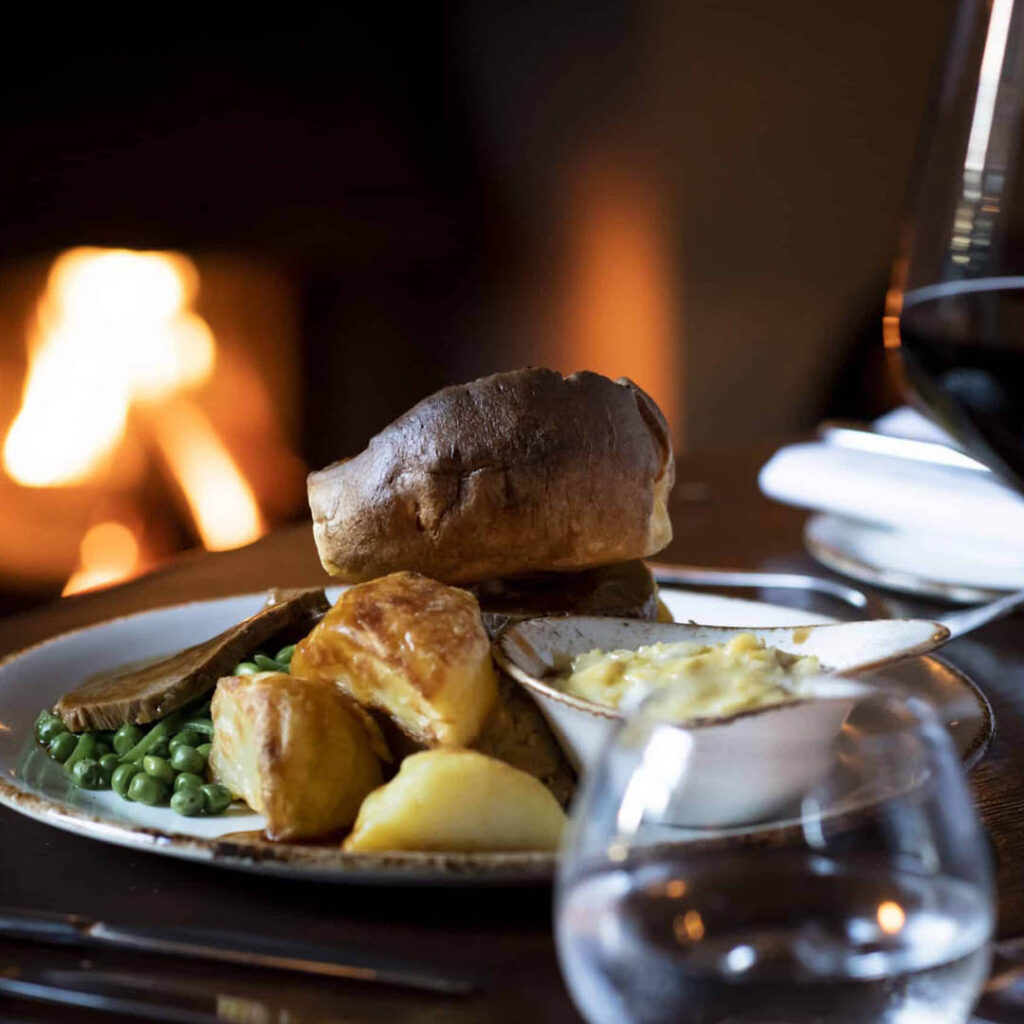 A plate of sliced beef, roast potatoes, peas and yorkshire puddings with gravy. A fire flickers in the background.