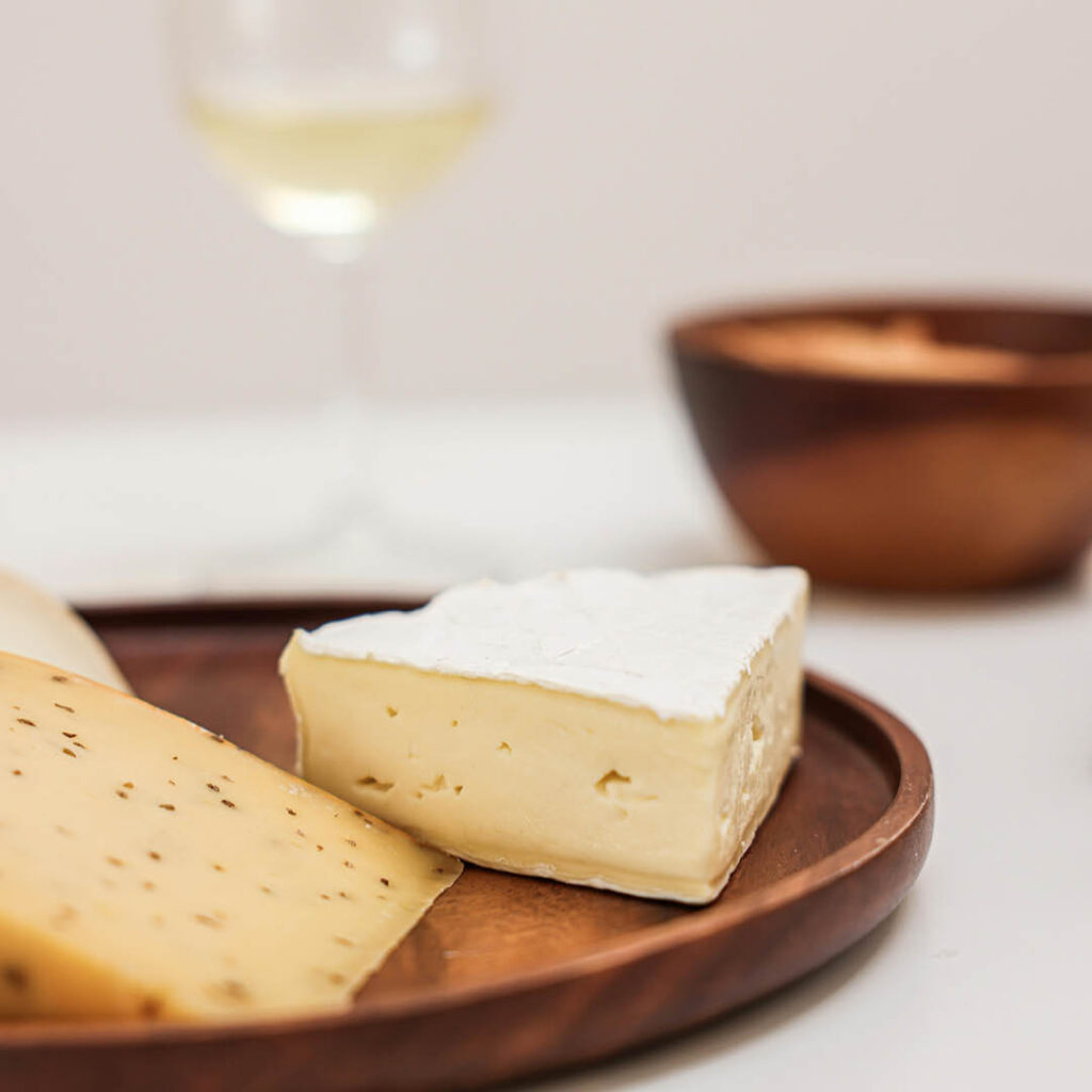 Two wedges of cheese sit on a brown wooden plate. A glass of white wine can be seen in the background.