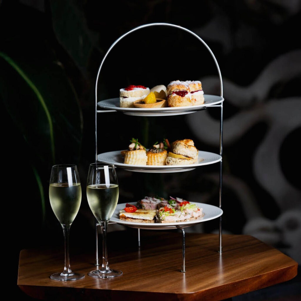 A three tier afternoon tea stand displaying dainty cakes, scones, sandwiches and other treats. Two glasses of prosecco stand beside it on the wooden table.