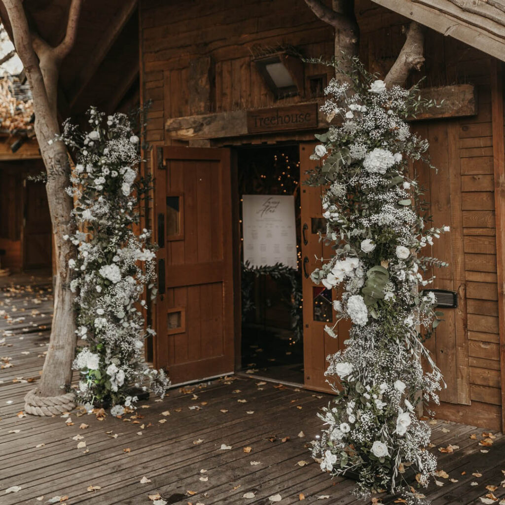 A wooden cabin with open doors is decorated on either side by white and green foliage. A wooden sign above the door reads 'Treehouse'. It is one of the wedding venue options at The Alnwick Garden.