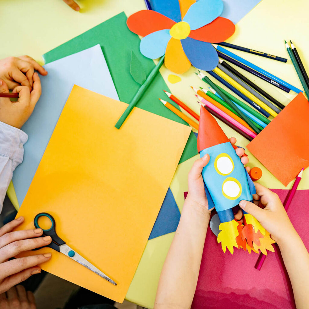 Unseen children play with multi-coloured sheets of paper. A paper flower lies flat on the table, while one child puts together a paper rocket.