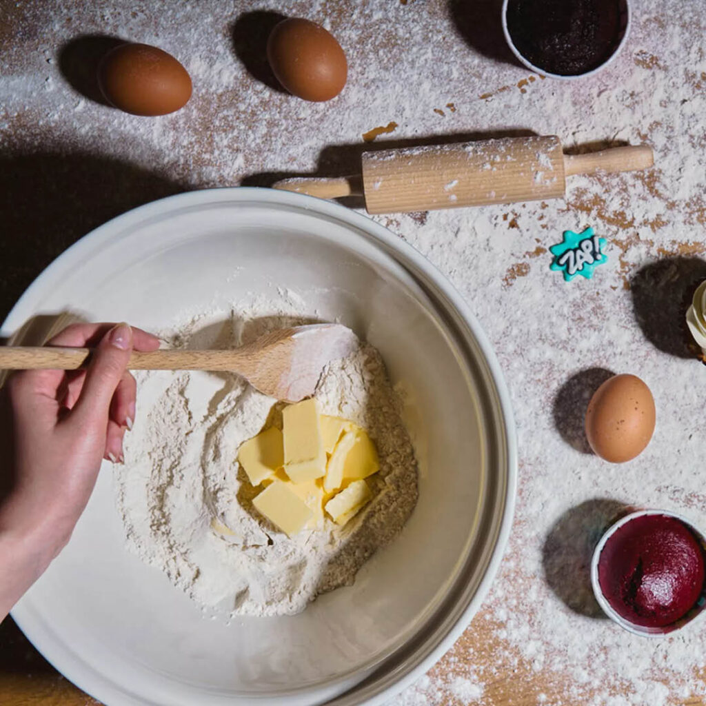 A person stirs butter and flour together in a bowl. The surface around the bowl is dusted with flour and scattered with eggs, food colouring and a wooden rolling pin.