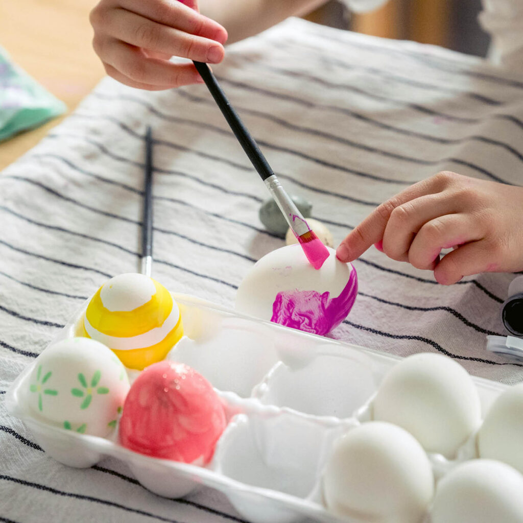 An unseen child carefully paints a white egg with purple paint. Already painted eggs sit in a white egg box.