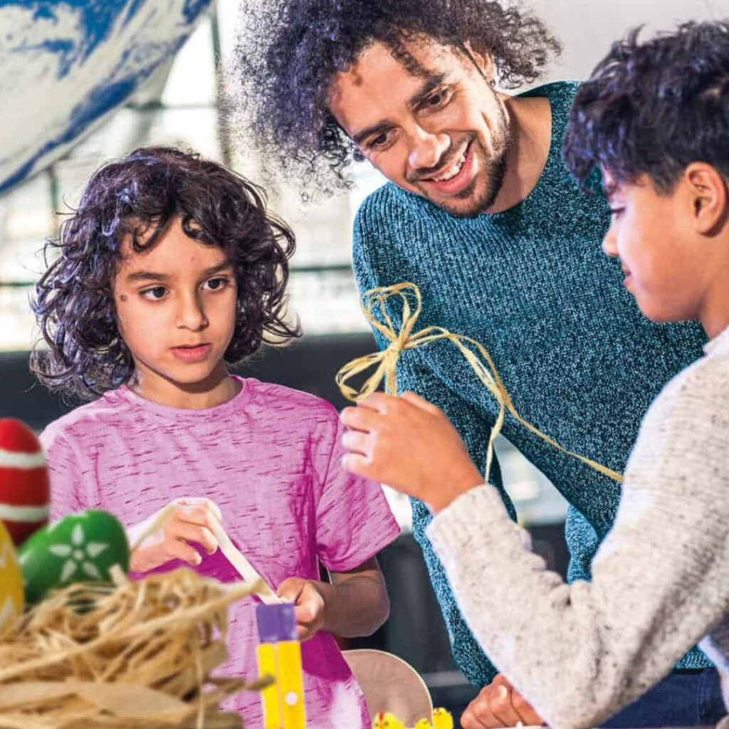 A father smiles as he watches his son tie twine into a bow shape. A young girl, maybe his sister, looks on in interest. A basket of colourful Easter eggs sits in front of them.