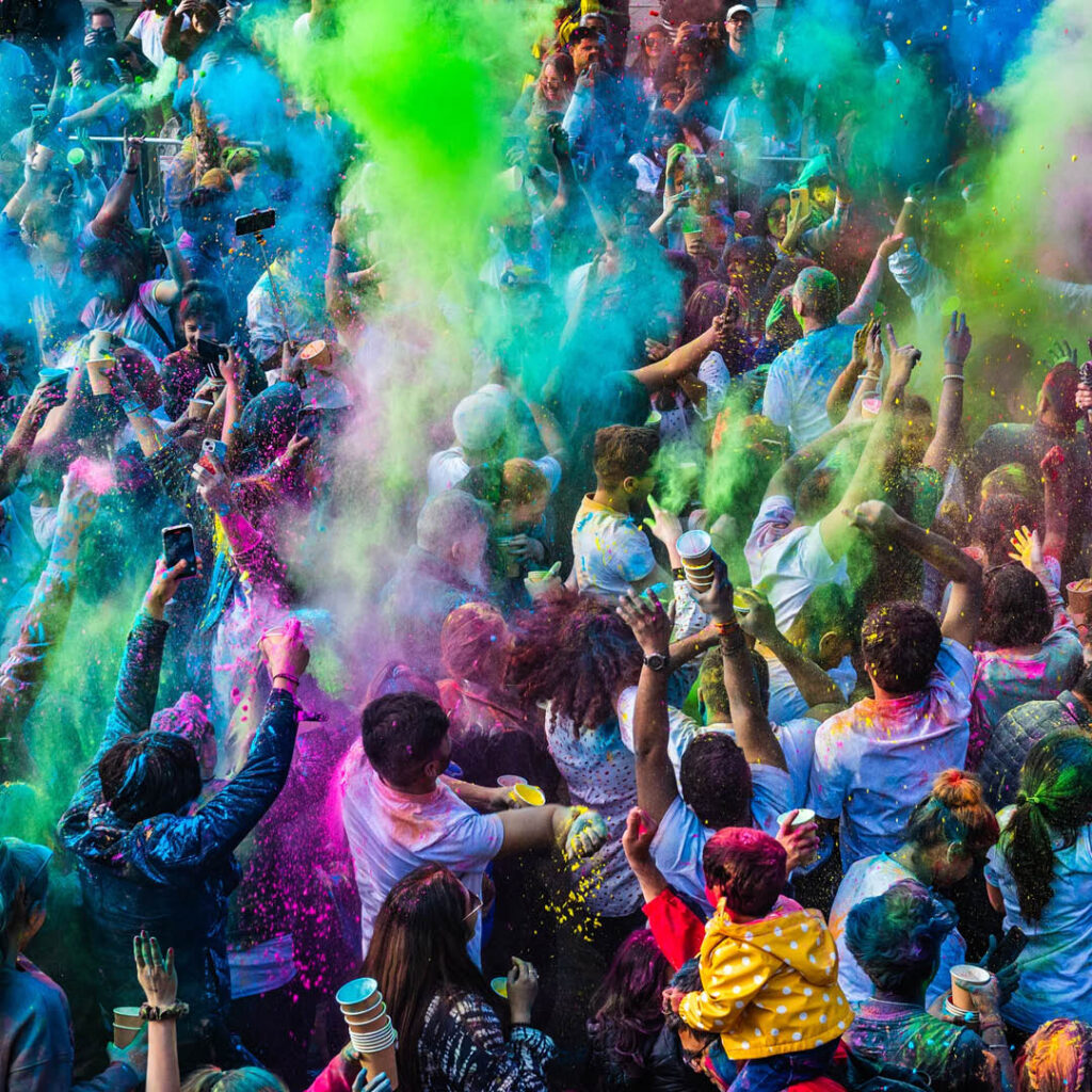 Aerial view of a crowd of people. They raise their hands in the air as clouds of coloured pigment float through the air and coat their skin. They are enjoying the colour throwing event of the Holi Festival at BALTIC.