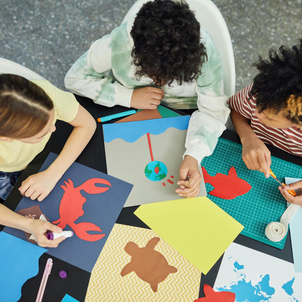 Three children sit around a table covered in coloured paper and cut outs of animals. They lean over as they stick the animals to sheets of the paper.