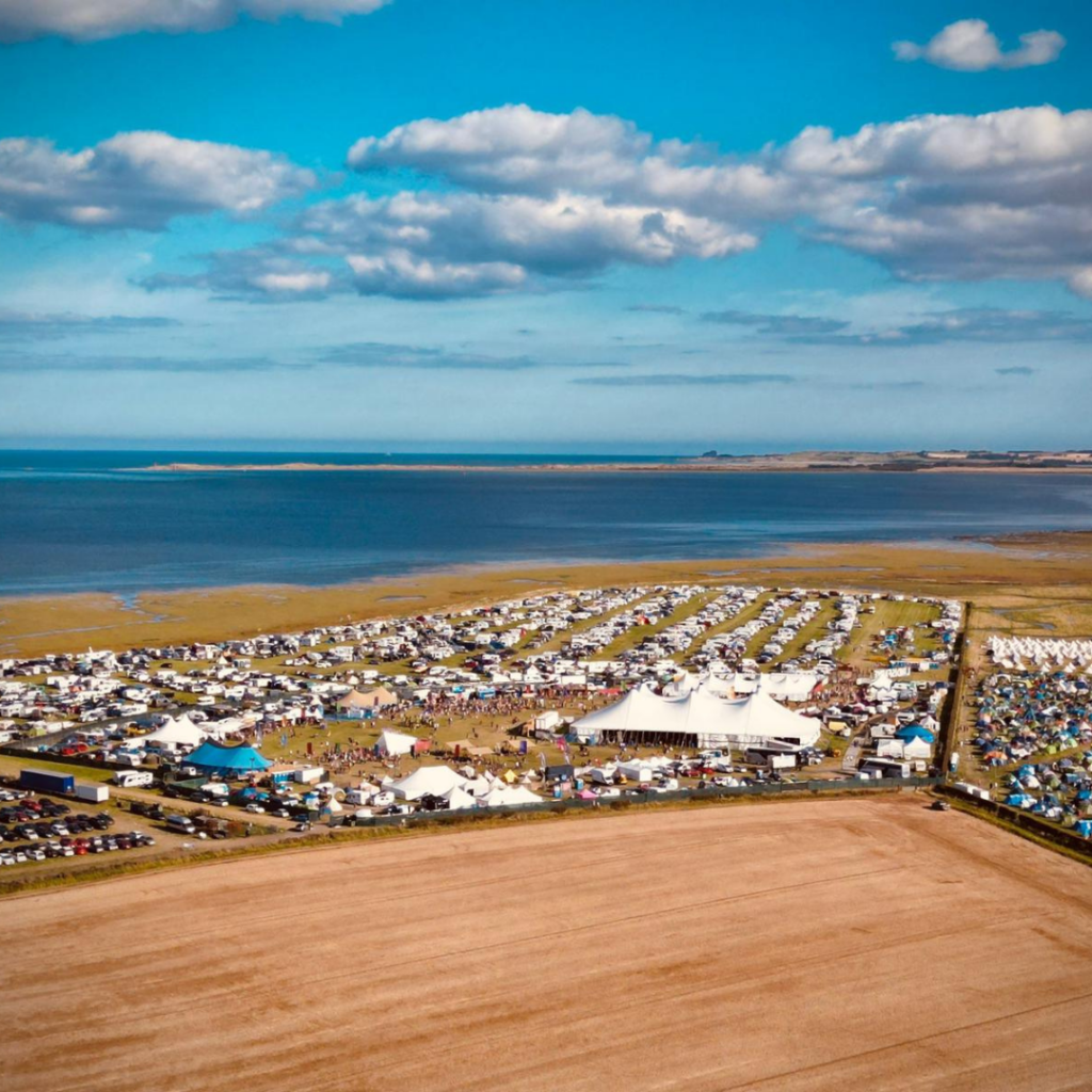 Aerial view of the Lindisfarne Festival. Tents, gazebos and crowds of people scatter across a sunny clifftop overlooking blue sea.