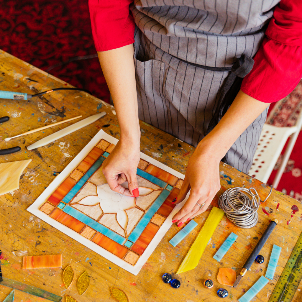 A person in a grey striped apron carefully places a small glass piece on a mosaic tile.