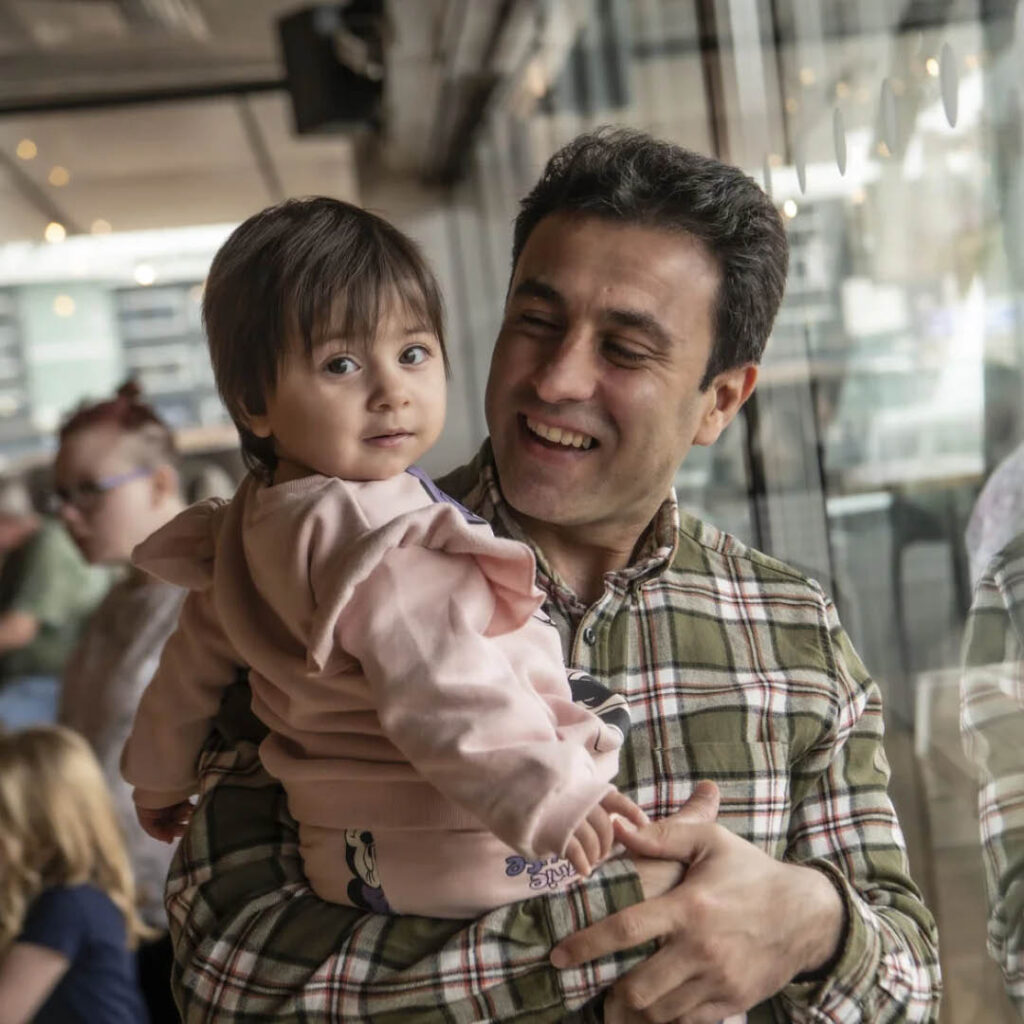 A man in a checkered green shirt grins as he looks at his toddler daughter, who he holds in his arms. The little girl twists around to look at the camera with a small curious smile on her face.