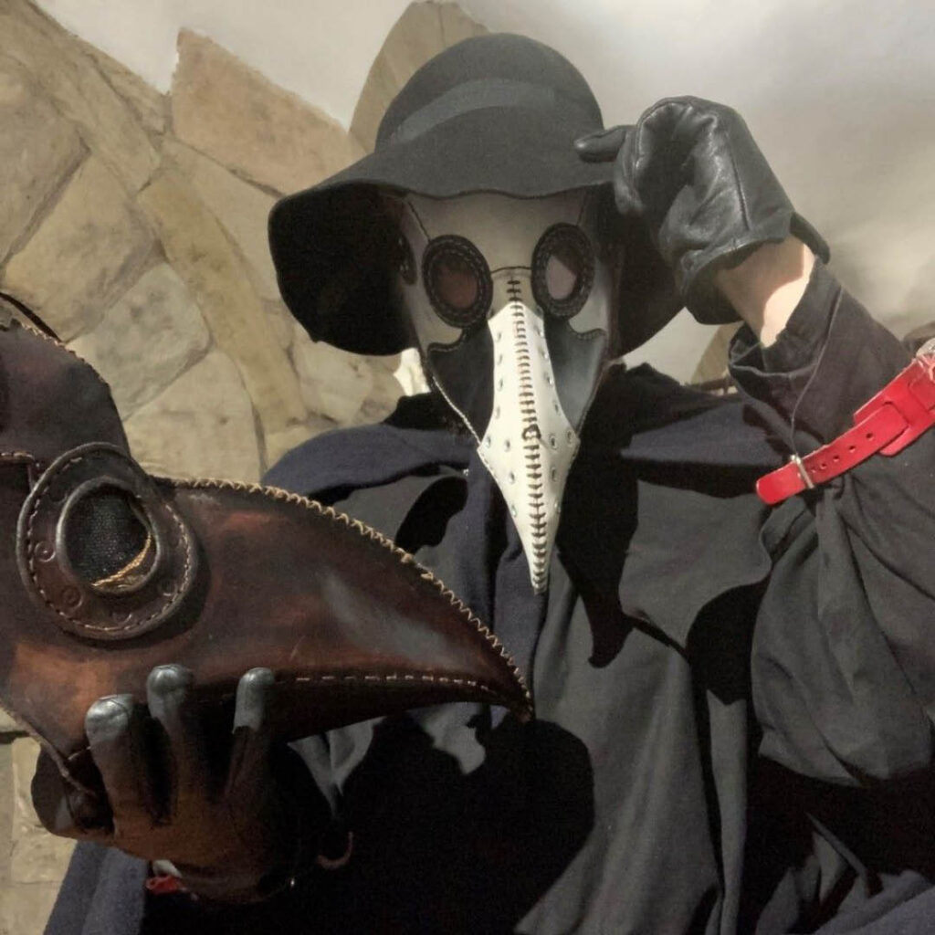 A person dressed as a plague doctor tips their black hat at the camera. In their gloved hand, they hold another brown leather plague mask.