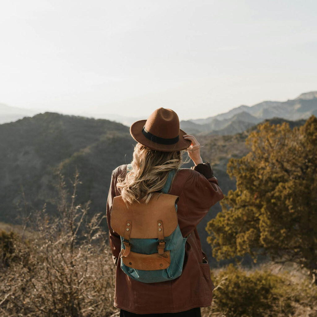 A woman wearing a brown hat, coat and yellow rucksack looks out at a mountainous landscape beyond.