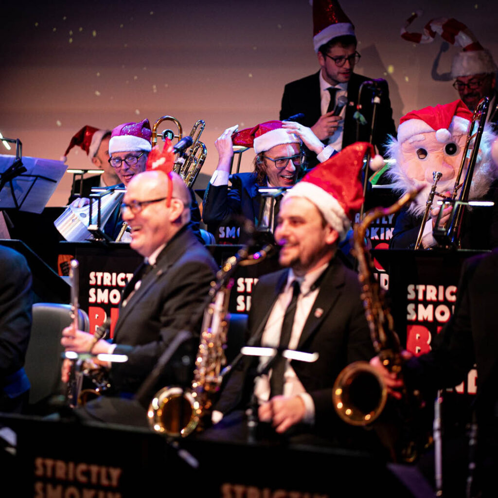 Musicians in the Strictly Smokin Big Band sit on stage. They wear Santa hats and laugh as they hold their various instruments, including flutes, saxophones and trombones.