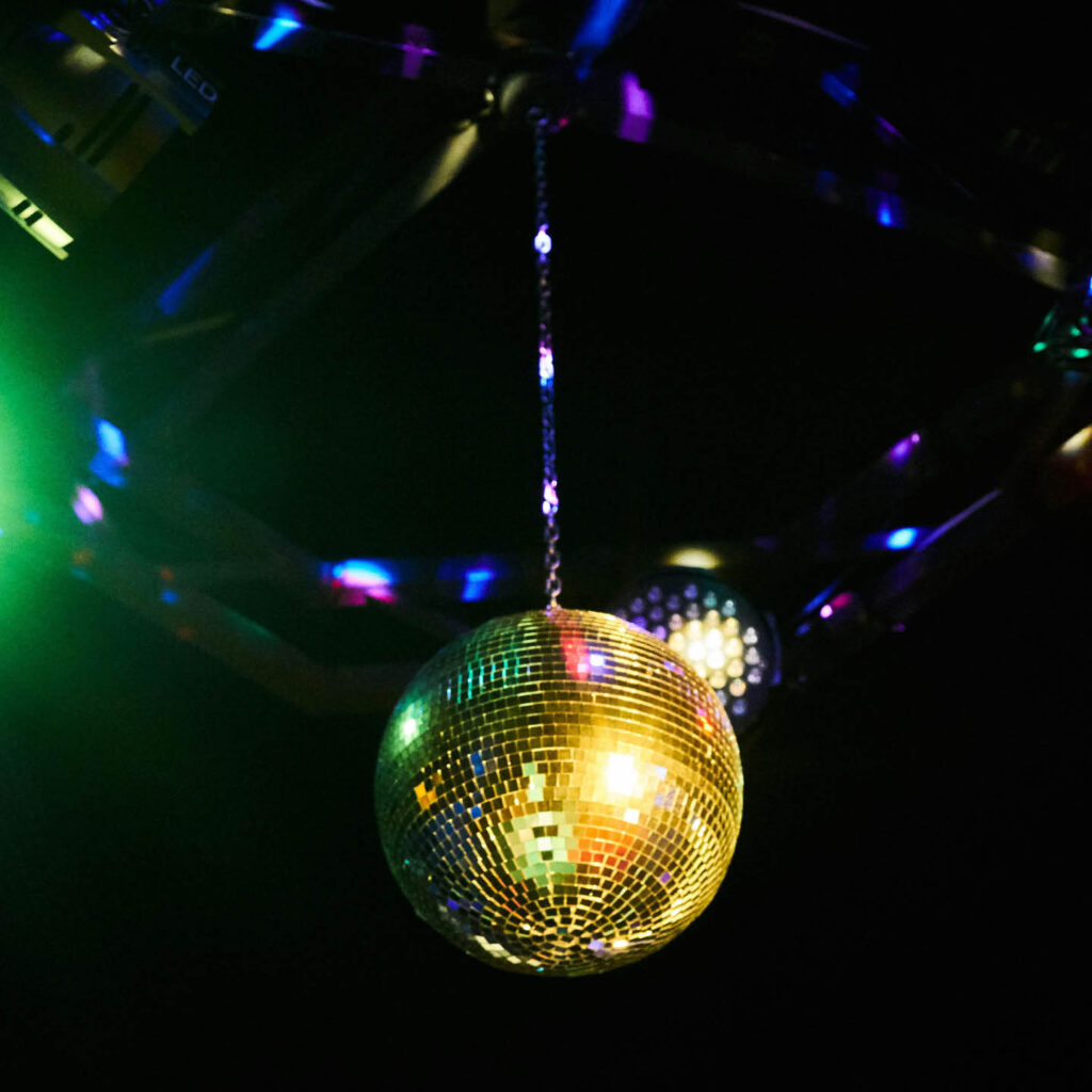 A large disco ball hangs from a black ceiling. Multi coloured shards of light glimmer around it.