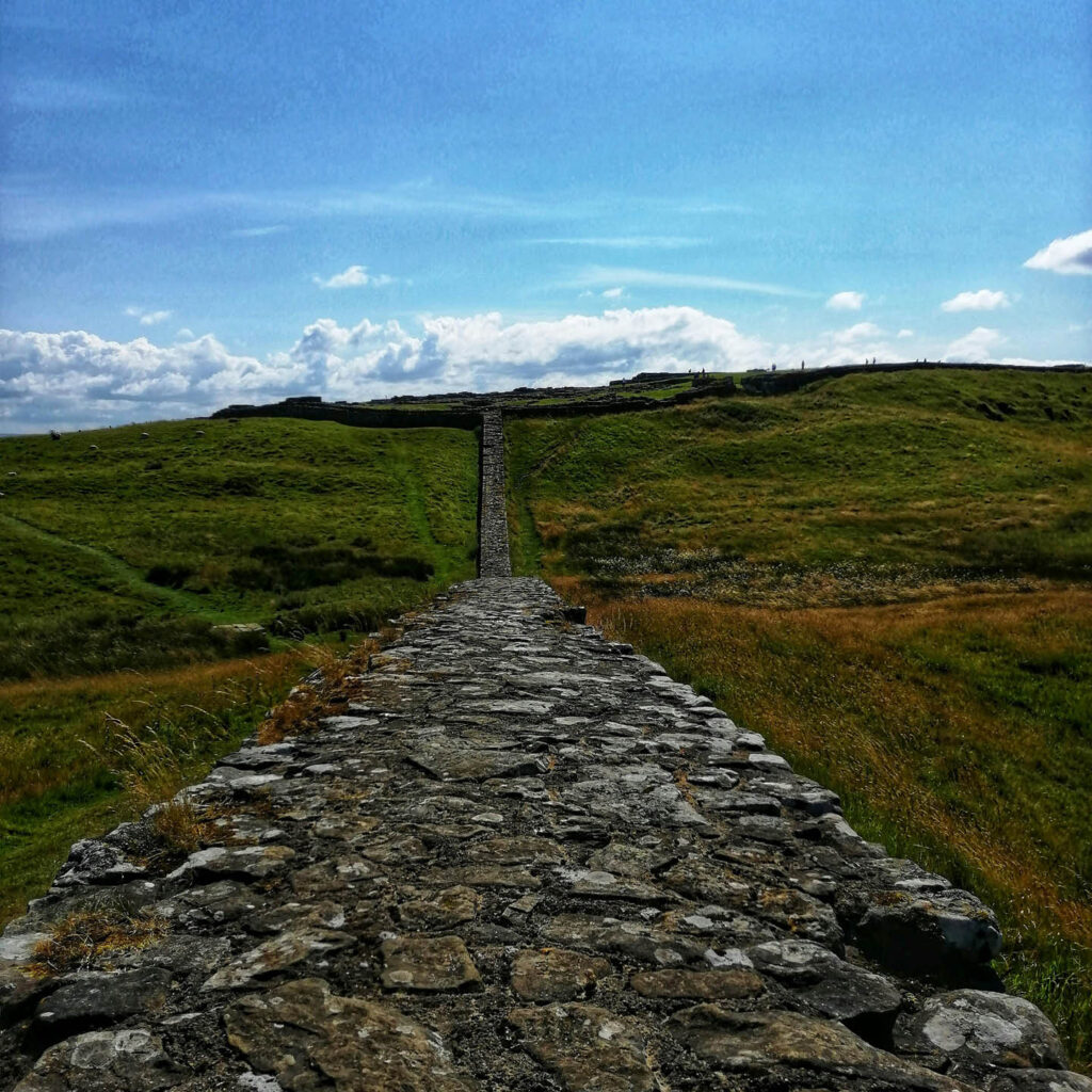 Hadrian's Wall stretches towards the horizon beneath a bright blue sky. Green fields fall away from it on both sides.