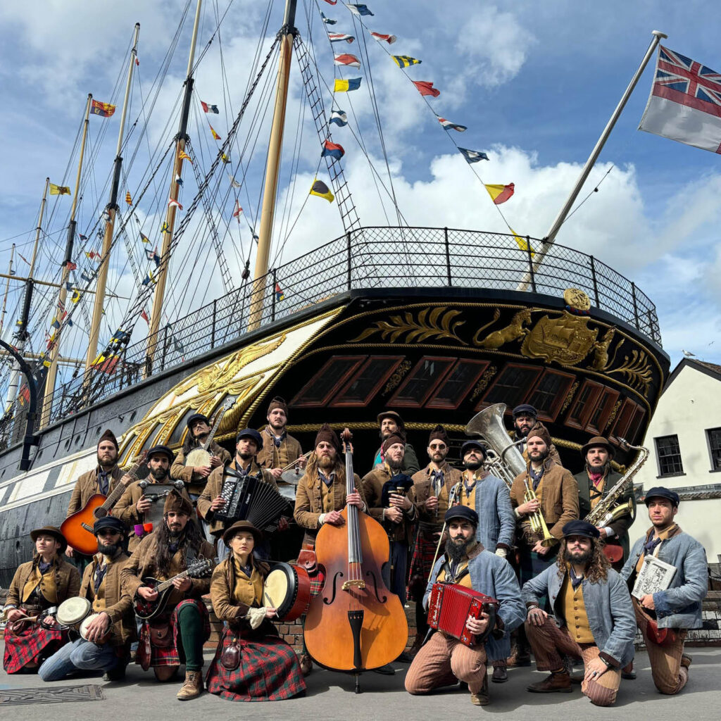 A big group of people wearing various forms of pirate or sailor outfits sit and stand in front of an old ship. They hold a range of instruments, from string to brass.