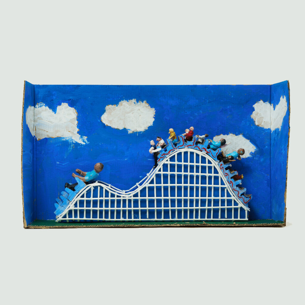 A cardboard stage has been painted deep blue with white clouds. A sculpture of a rollercoaster has been built inside it. It features different figurines of people seated inside two rollercoaster trains. They travel over two bumps.
