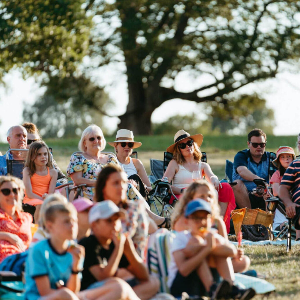 People wearing sunglasses and sun hats sit on the grass in Raby Castle's grounds. Sun shines down as they watch an unseen amphitheatre show.