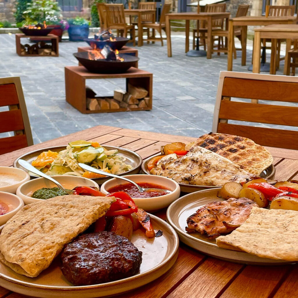 Various plates of BBQ meats, pitta bread, salad and grilled vegetables. They are laid on a wooden table in the cloister garden of Blackfriars Restaurant. A fire pit burns in the background.