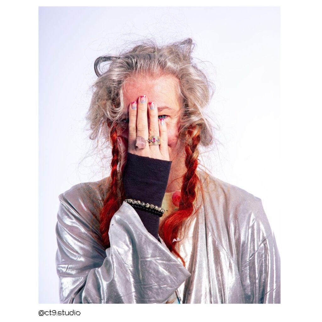 Delaine Le Bas is a white woman with unruly grey hair plaited into two red pigtails. She wears a silver robe over a black. She covers her face with one hand, one eye peeking out playfully.