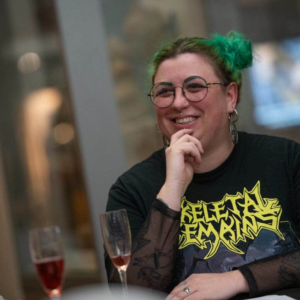 A woman with green hair pulled into space buns grins as she sits at a table at one of the Discovery Museum's Discovery Nights. An almost empty glass of fizz sits in front of her.