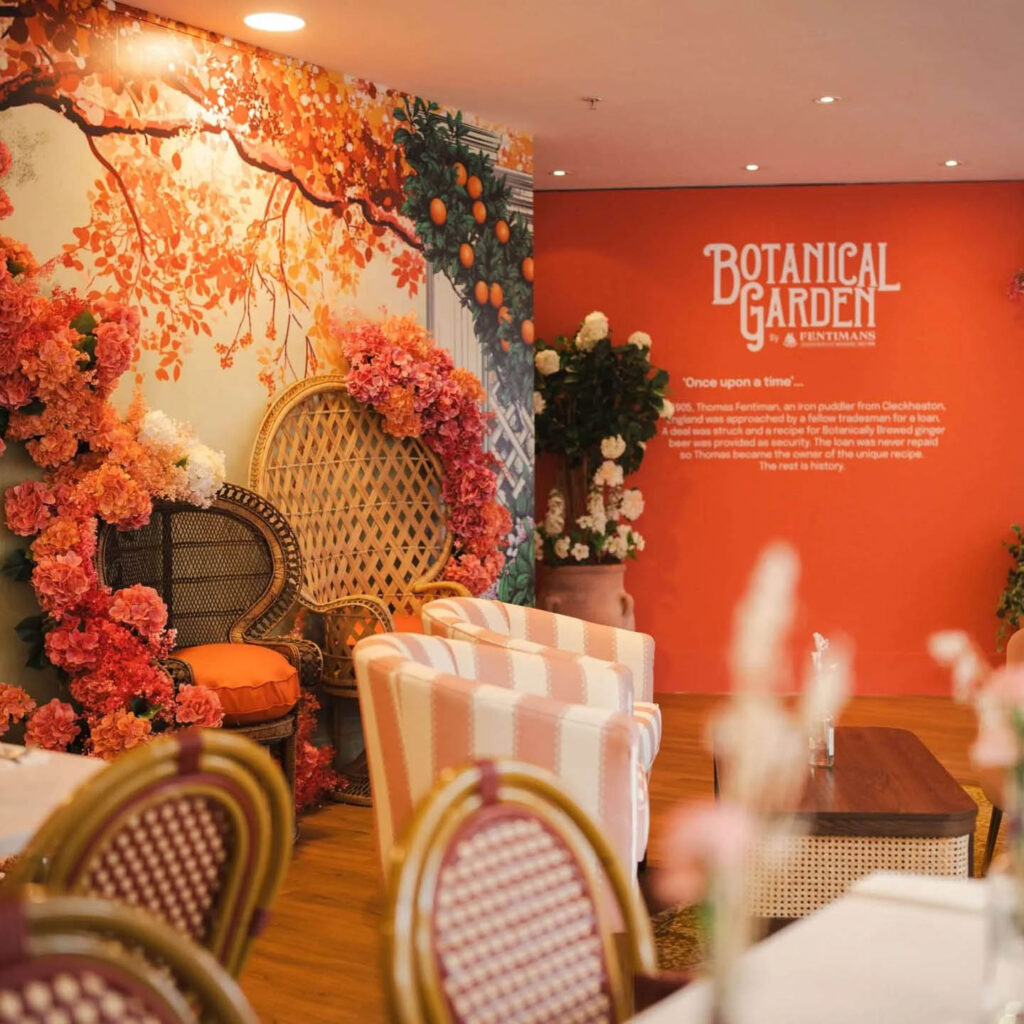 A dining space inside Fenwick Newcastle. The walls are orange with paintings of orange trees. Orange and white striped armchairs and wicker backed chairs are placed around tables. This is the garden room - part of Fenwick and Fentimans collaboration.