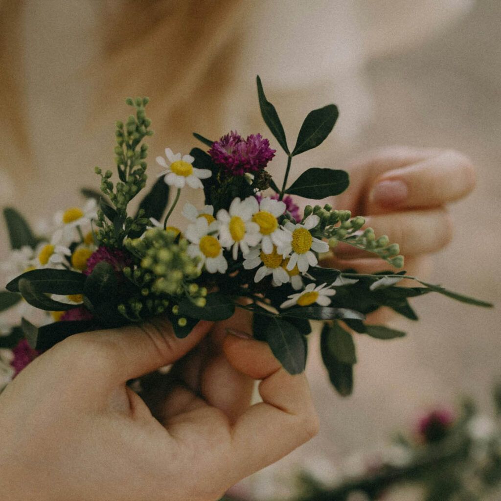 A person holds a delicate flower wreath in their hands. It is made from green leaves, daisies and purple flowers.