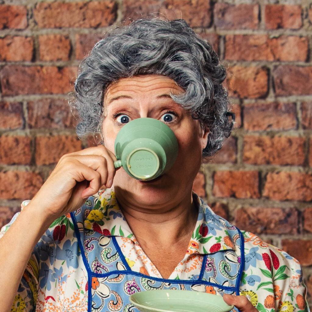 A woman wearing a grey perm wig raises her eyebrows as she drinks from a green tea cup. She wears a colourful floral shirt and apron. She is one of the Two Lucys in Housewife at War.